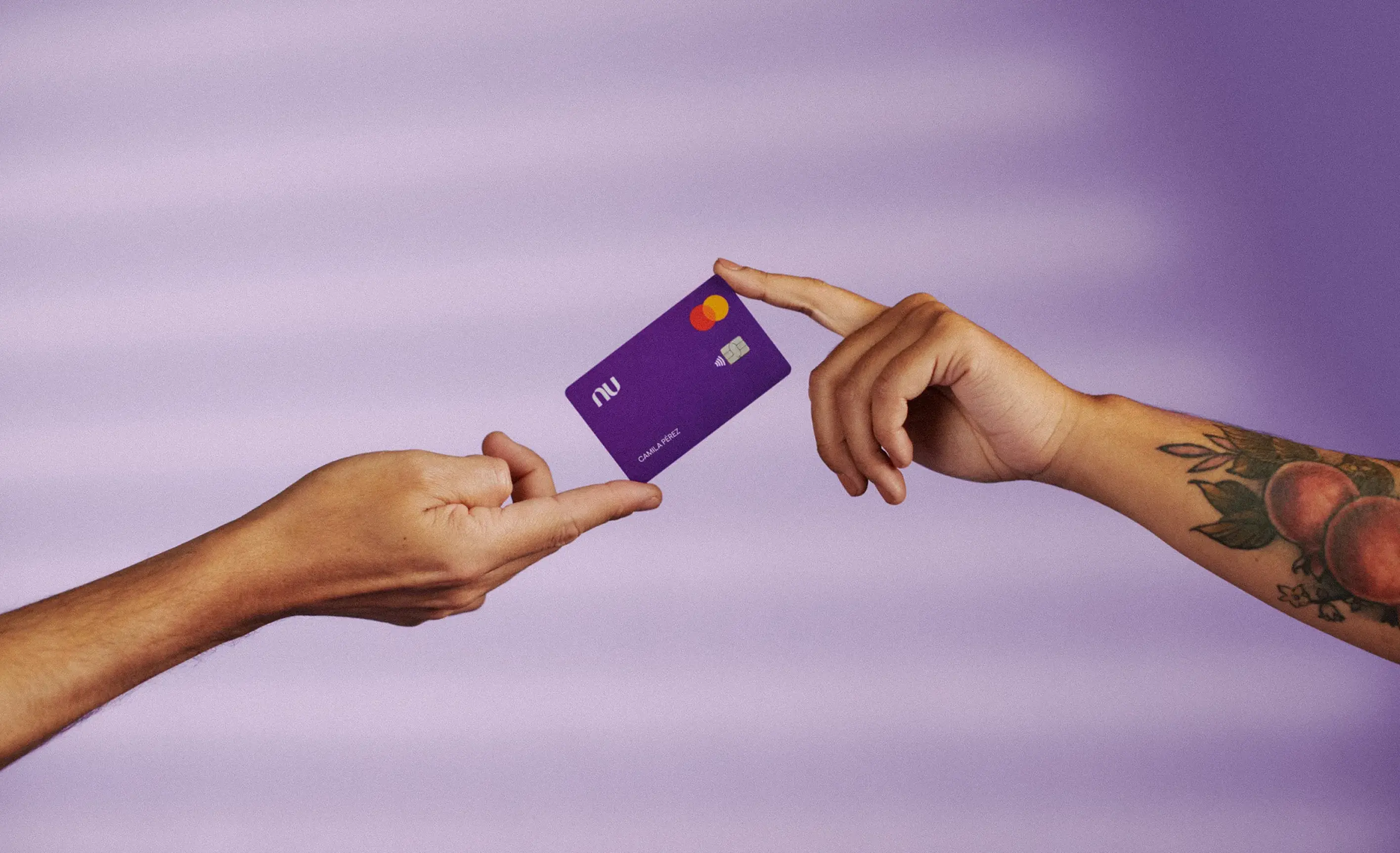 Two hands against a purple background exchanging a purple NU credit card, one hand has a tattoo with peaches and leaves.