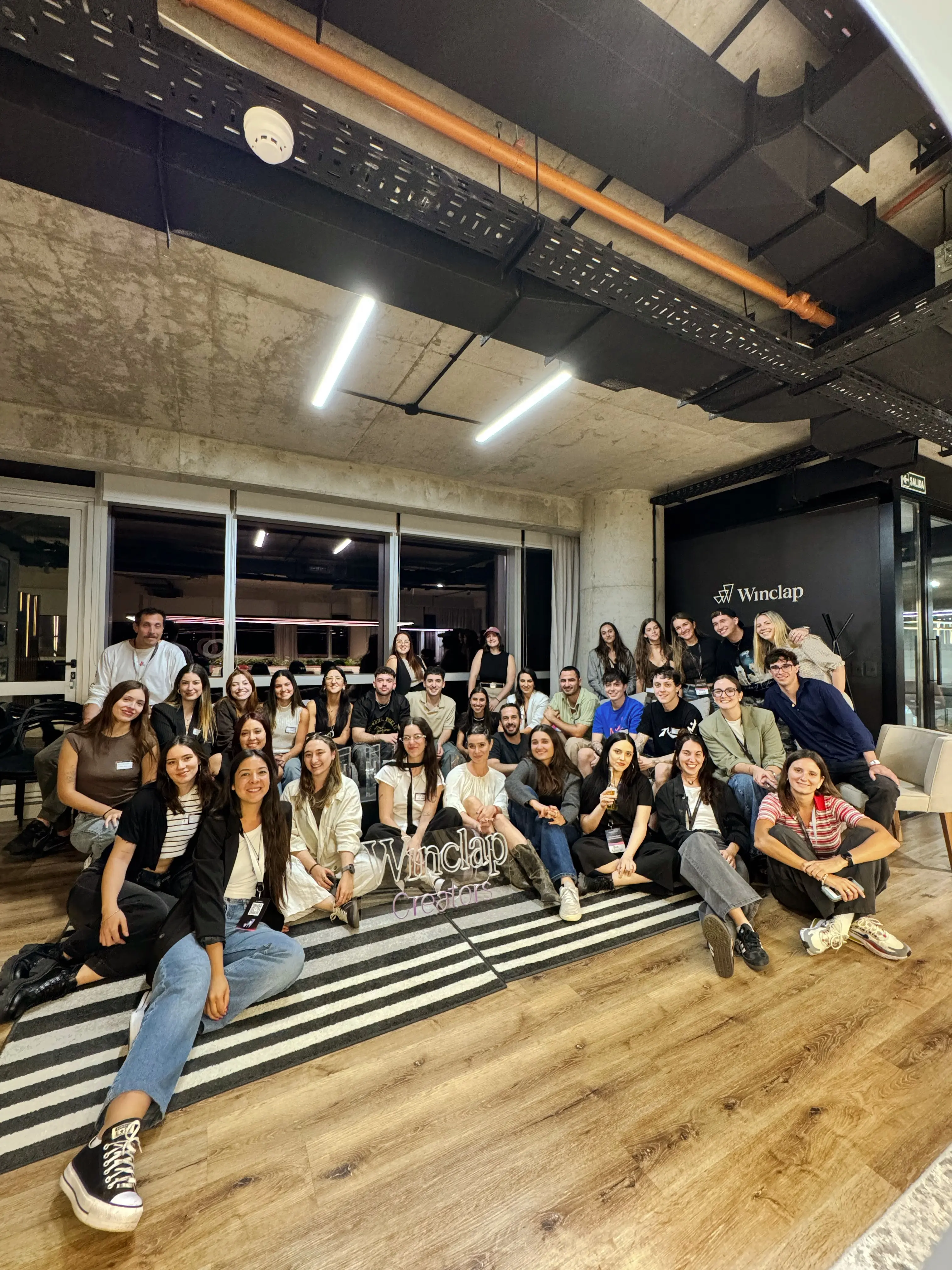 Group of about 30 diverse young adults seated and standing indoors in a brightly lit office space with a Winclap sign on the wall.