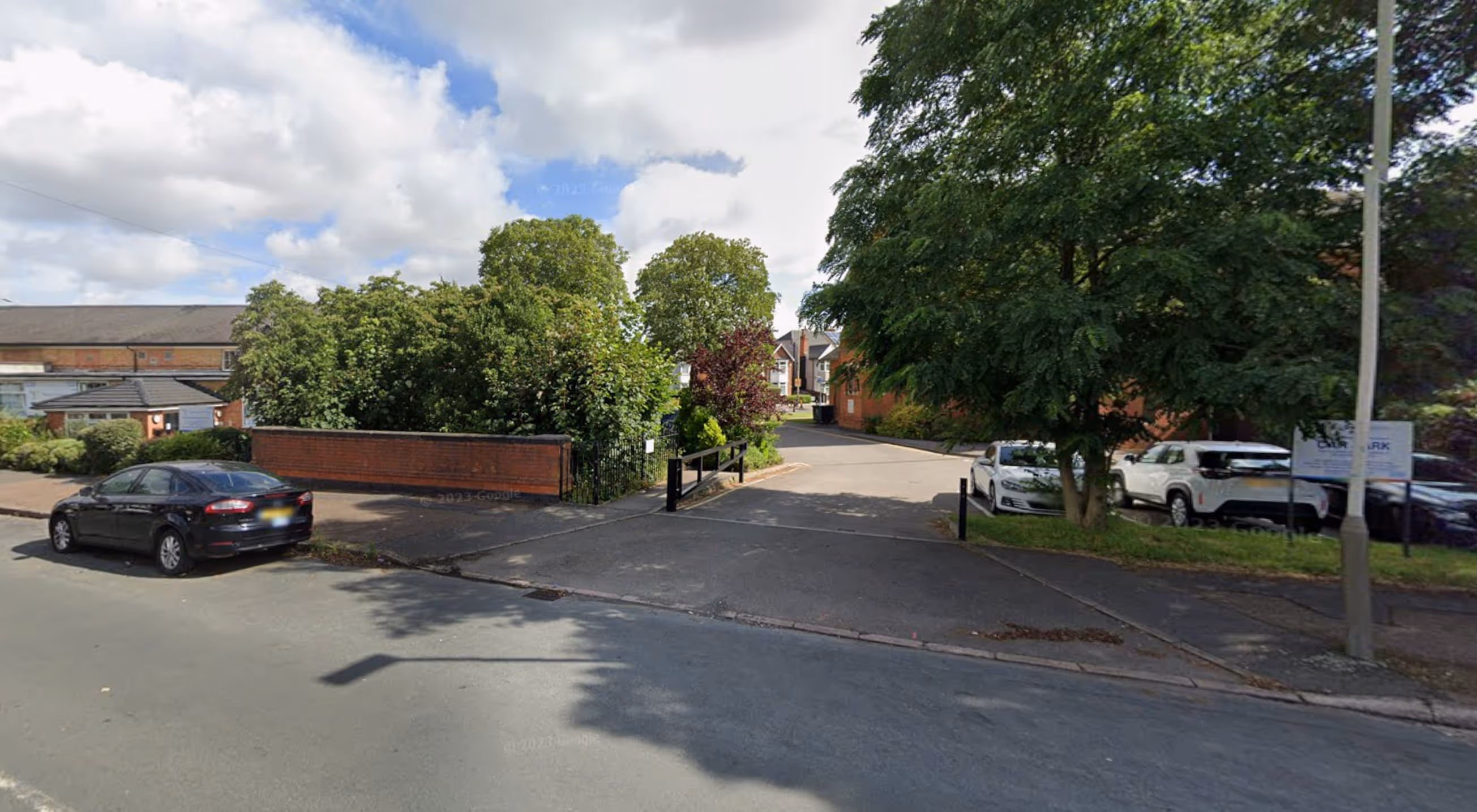 Residential street entrance with parked cars, trees, and a partly cloudy sky.
