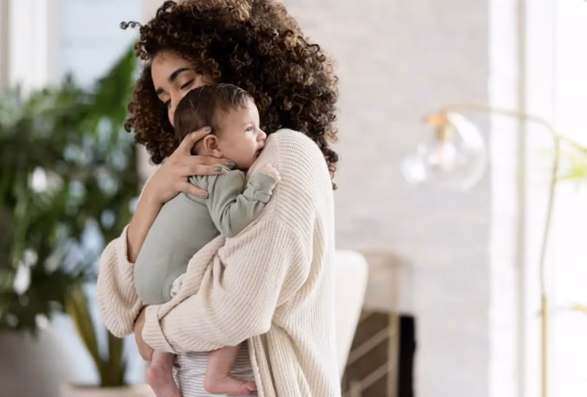Woman with curly hair gently holding and hugging a baby indoors near a plant and light fixture.