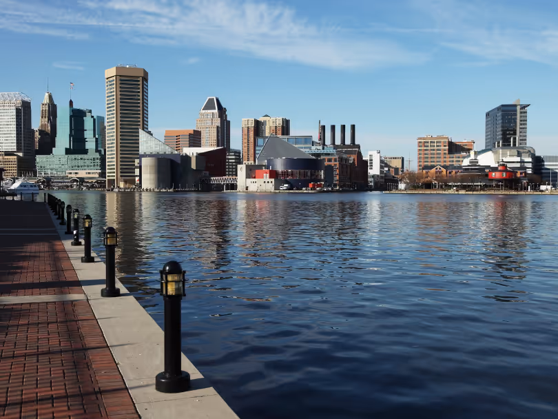 Waterfront promenade with lamp posts and a city skyline of modern buildings across the calm harbor under a partly cloudy blue sky.