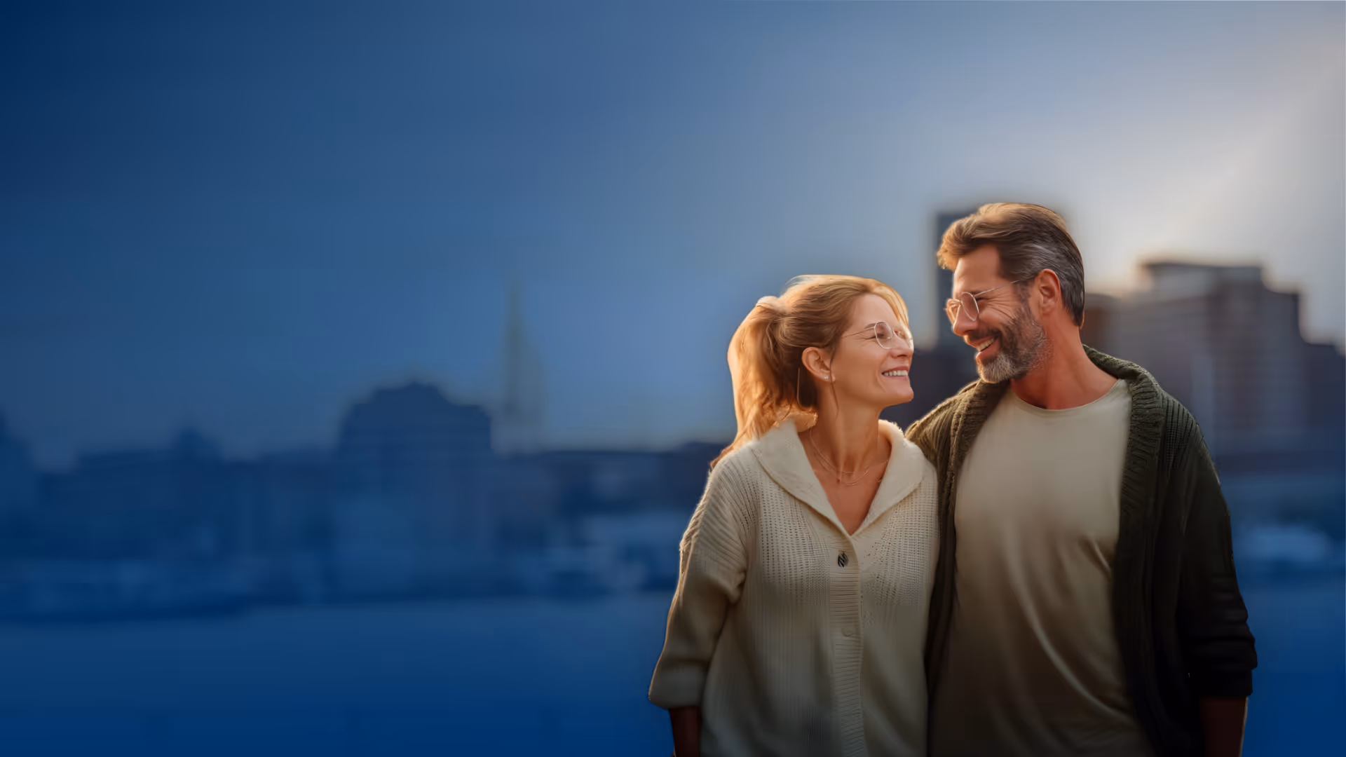 Smiling middle-aged couple wearing glasses embracing outdoors with city skyline blurred in the background.