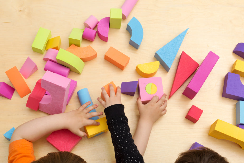Two children’s hands playing with colorful geometric wooden blocks on a light wood surface.