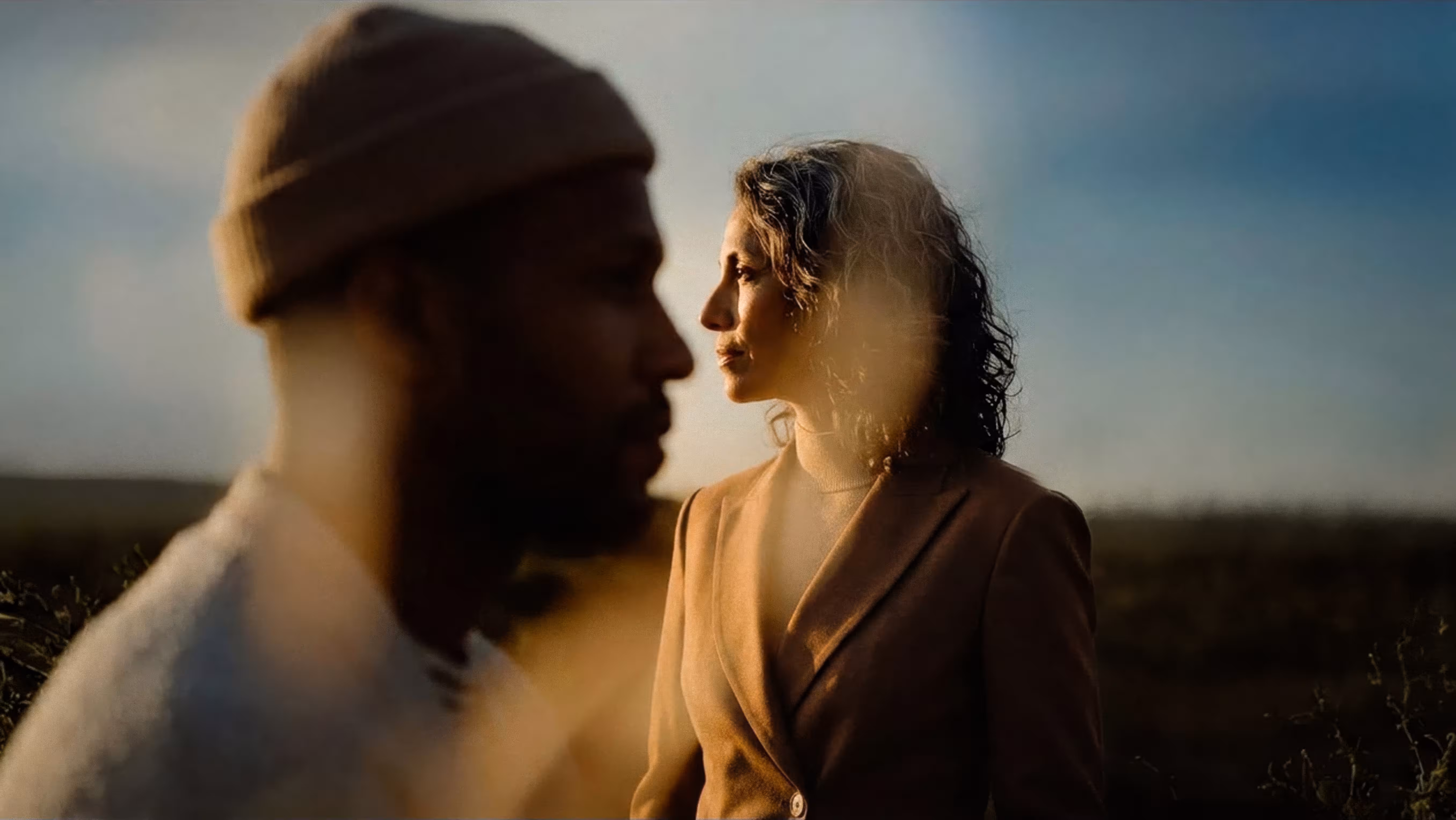 Profile of a woman with curly hair wearing a brown coat in soft light, with a blurred man in a beanie in the foreground.