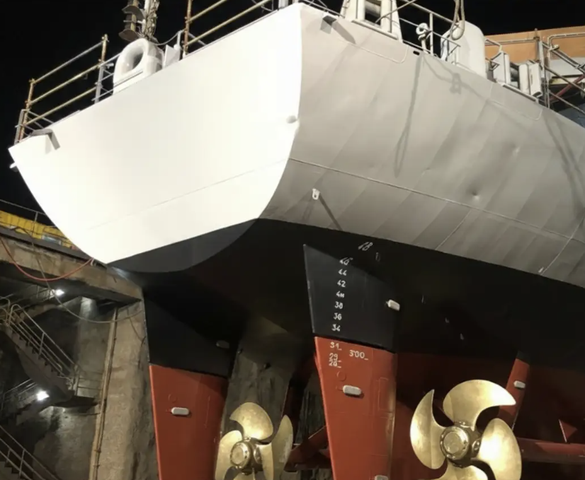 Vue de la poupe d'un navire militaire dans une cale sèche, montrant ses deux hélices dorées et sa coque blanche.