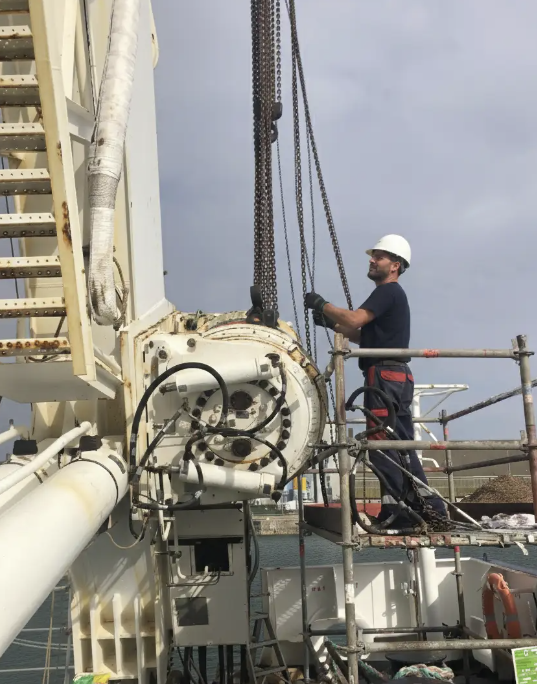 Ouvrier portant un casque de sécurité manipulant des chaînes sur une plateforme de travail en hauteur près d'une structure industrielle blanche au bord de l'eau.