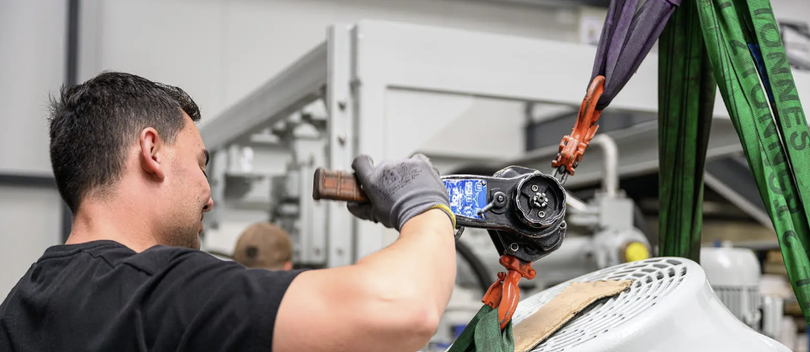 Un ouvrier utilisant un cric à cliquet pour ajuster une charge suspendue avec des sangles dans un environnement industriel.