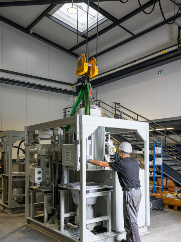 Un ouvrier portant un casque de sécurité travaille sur une grande machine industrielle suspendue par une grue à l'intérieur d'un atelier.