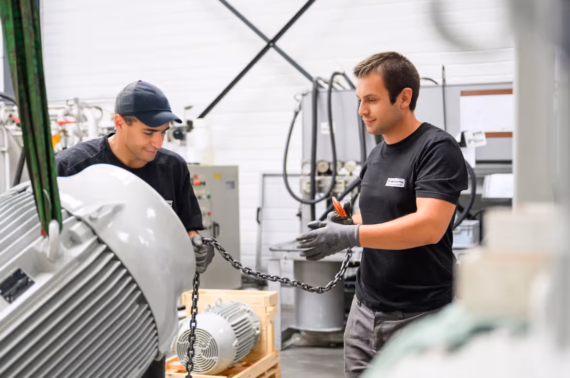 Deux techniciens en gants gris manipulent une chaîne autour d'un gros moteur industriel dans un atelier.