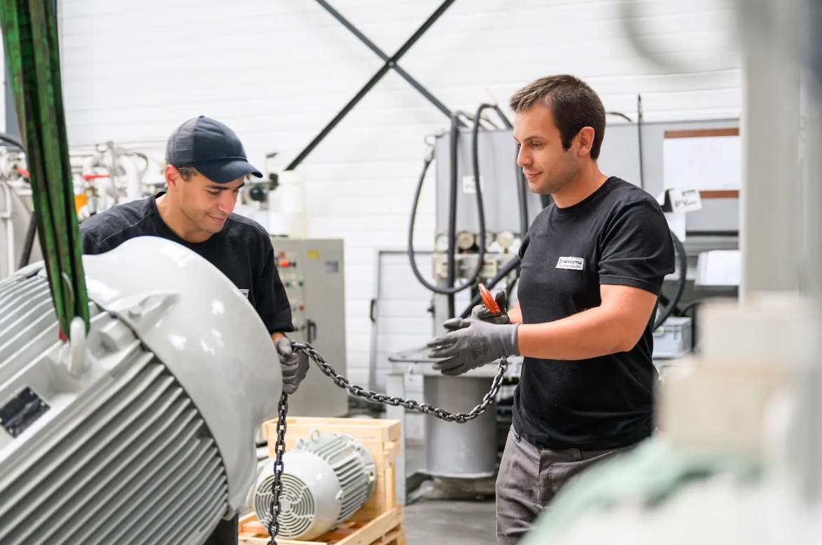 Deux techniciens en gants gris manipulent une chaîne autour d'un gros moteur industriel dans un atelier.