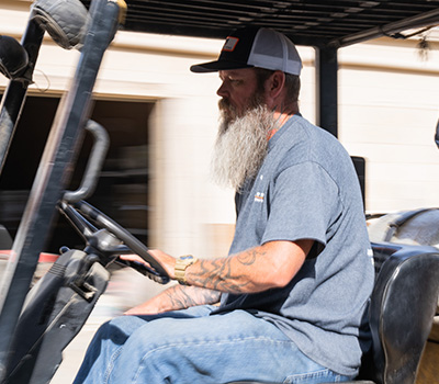 Man with a long white beard, tattooed arm, and a baseball cap driving a golf cart outdoors.