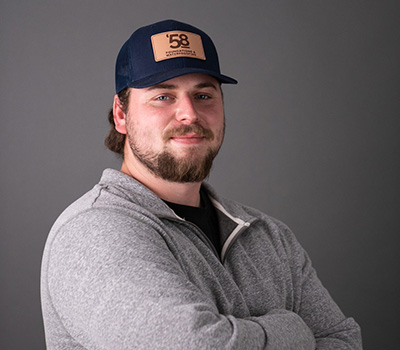 Bearded man wearing a navy blue cap and gray zip-up sweater, standing with arms crossed against a gray background.