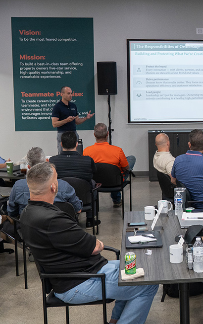 Presenter speaking in a meeting room to an audience seated around tables and chairs, with a slide presentation and a vision, mission, and team promise poster on the wall.