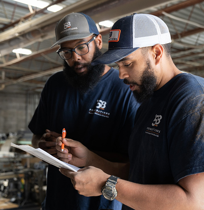 Two men in 58 Foundations shirts and caps reviewing papers indoors in an industrial setting.
