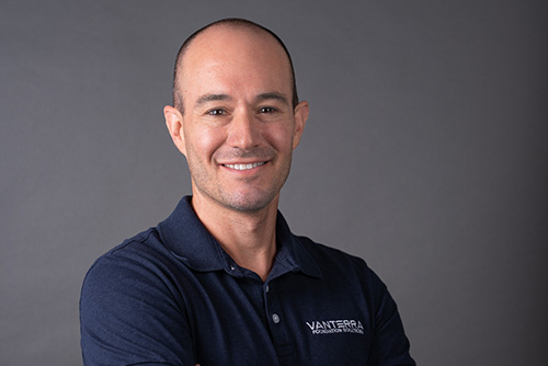 Smiling bald man wearing a navy blue Vantara polo shirt with arms crossed against a gray background.