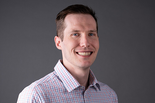 Smiling man with short brown hair wearing a checkered shirt against a plain gray background.
