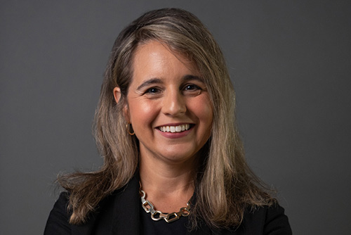 Smiling woman with shoulder-length light brown hair wearing a black blazer and gold chain necklace against a gray background.