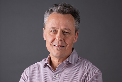 Middle-aged man with gray hair wearing a striped button-up shirt against a gray background.