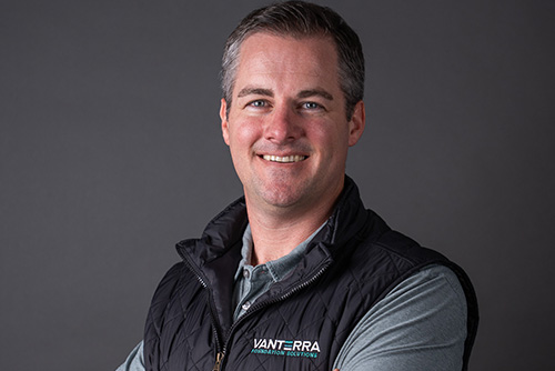 Smiling man with short hair wearing a black VANTERRA Innovation Ecosystems vest over a gray shirt against a gray background.