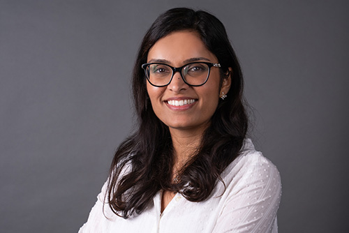 Smiling woman with glasses and long dark hair wearing a white blouse against a gray background.
