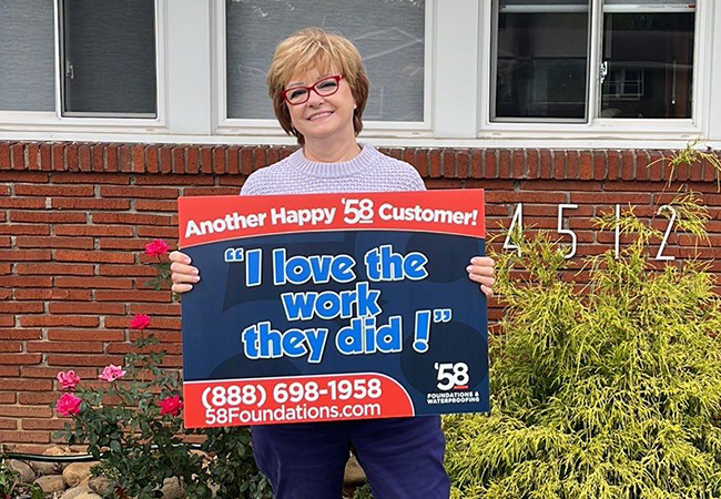 Smiling woman with short blonde hair and red glasses holding a sign that says 'Another Happy 58 Customer! I love the work they did!' in front of a brick house with plants and house number 4512.