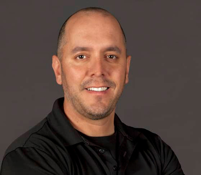 Smiling bald man with light beard wearing a black collared shirt against a dark gray background.