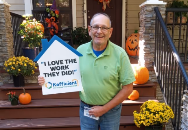 Smiling man in green shirt holding a house-shaped sign that says 'I love the work they did!' with Kefficient logo, standing on a porch decorated with pumpkins and yellow flowers.