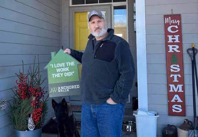 Man standing on a porch holding a green house-shaped sign that reads 'I LOVE THE WORK THEY DID!' with a black dog sitting beside him.
