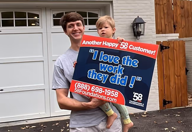 Smiling man holding a young child in front of a garage, both holding a sign saying 'Another Happy 58 Customer! I love the work they did!' with contact information for 58 Foundations & Waterproofing.