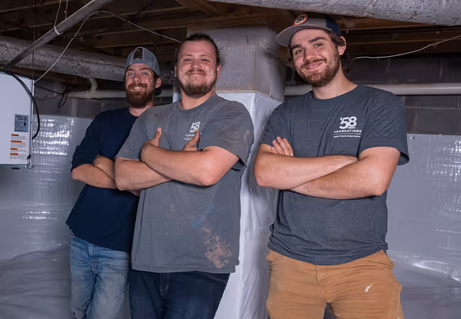 Three men wearing casual work clothes and smiling with arms crossed inside a basement under a wooden ceiling.