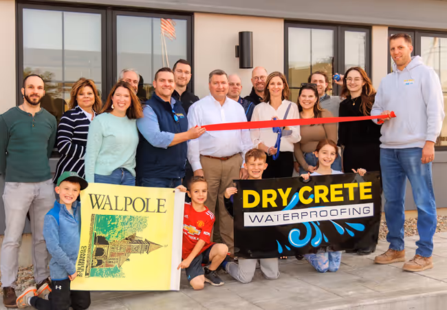 Group of adults and children standing outside a building holding a red ribbon and two signs reading Walpole and Dry Crete Waterproofing for a ribbon-cutting ceremony.