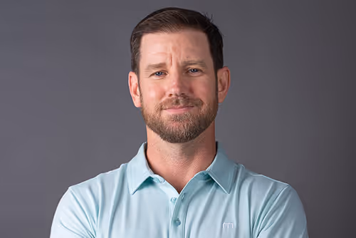 Will Portrait of a man with short brown hair and beard, wearing a light blue polo shirt, smiling against a gray background.