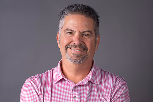 Todd a Middle-aged man with short salt-and-pepper hair and goatee smiling, wearing a pink checkered shirt against a gray background.