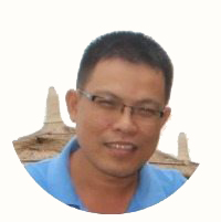 Man wearing glasses and a blue collared shirt smiling in front of a stone structure.