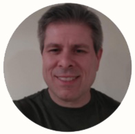 Smiling man with short gray hair wearing a dark shirt against a plain background.