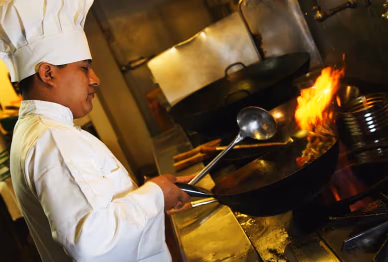 A chef in a white uniform and hat cooks with a wok, creating vibrant flames.