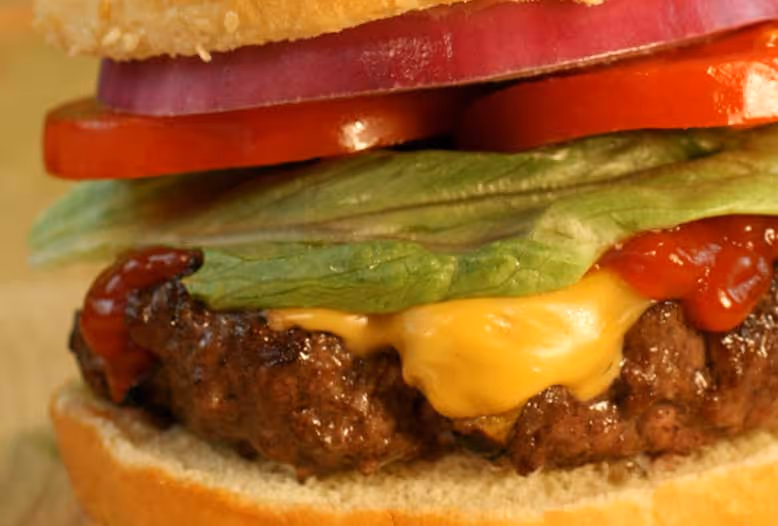 Close-up of a juicy cheeseburger with crispy lettuce, tomato slices, red onion, and melted cheese, topped with ketchup in a sesame seed bun.