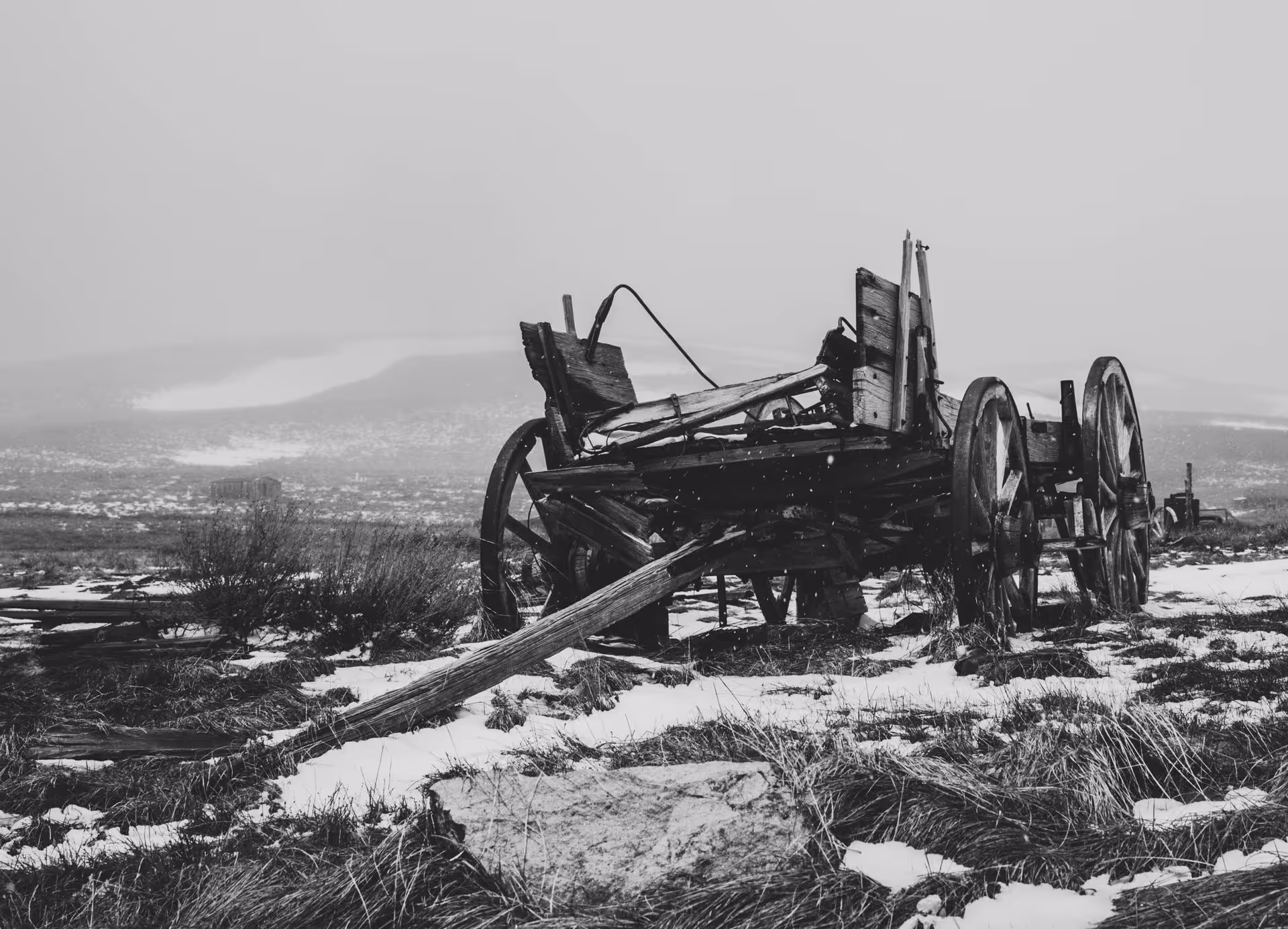 Black and white image of an old, broken wagon with large wooden wheels on a snowy, grassy landscape.