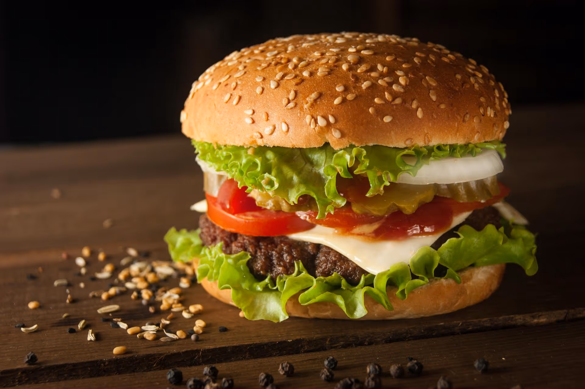 A juicy cheeseburger with a sesame seed bun, lettuce, tomato, onion, and pickles sits on a wooden table. Pepper and seeds are sprinkled around.