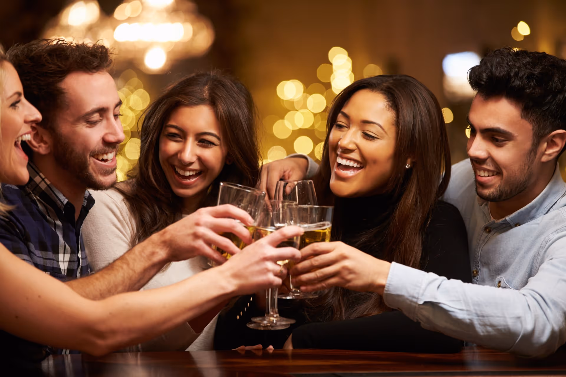 A group of five friends, three women and two men, joyfully clink glasses in a warmly lit setting, with soft golden lights in the background adding a festive tone.
