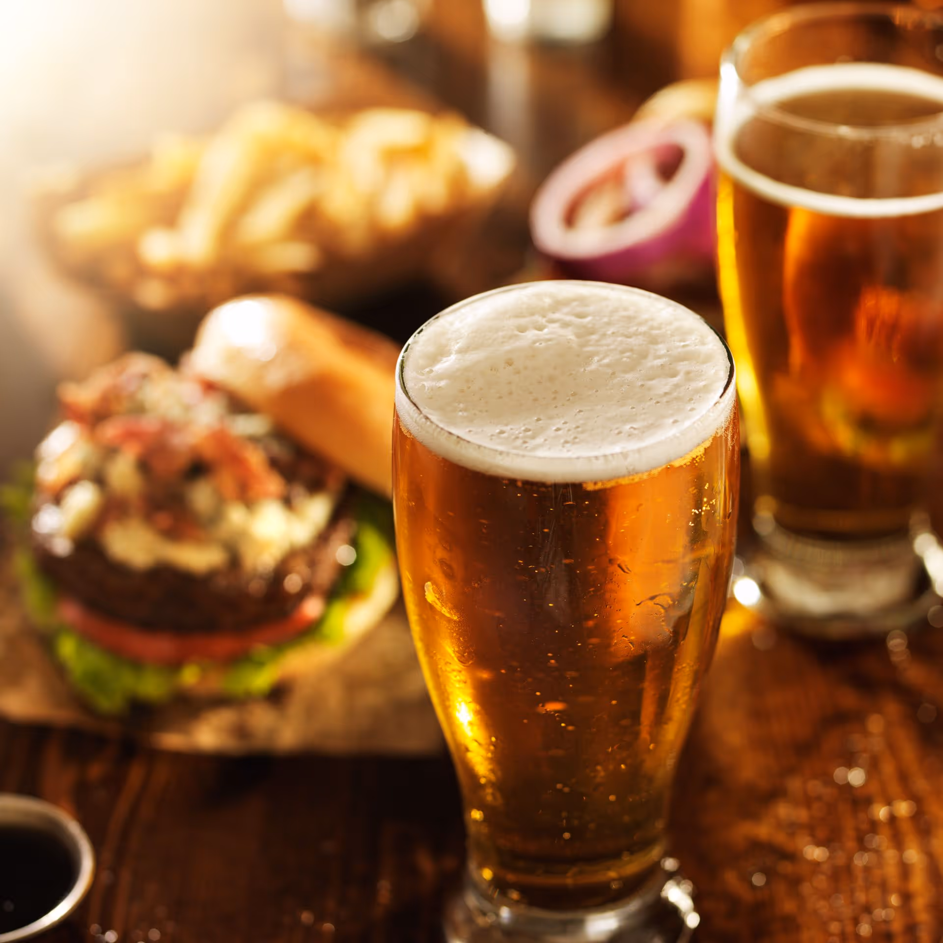Cold beer in a glass with frothy head on a wooden table, flanked by another beer, a juicy burger with toppings, and a bowl of golden fries. Warm, inviting ambiance.