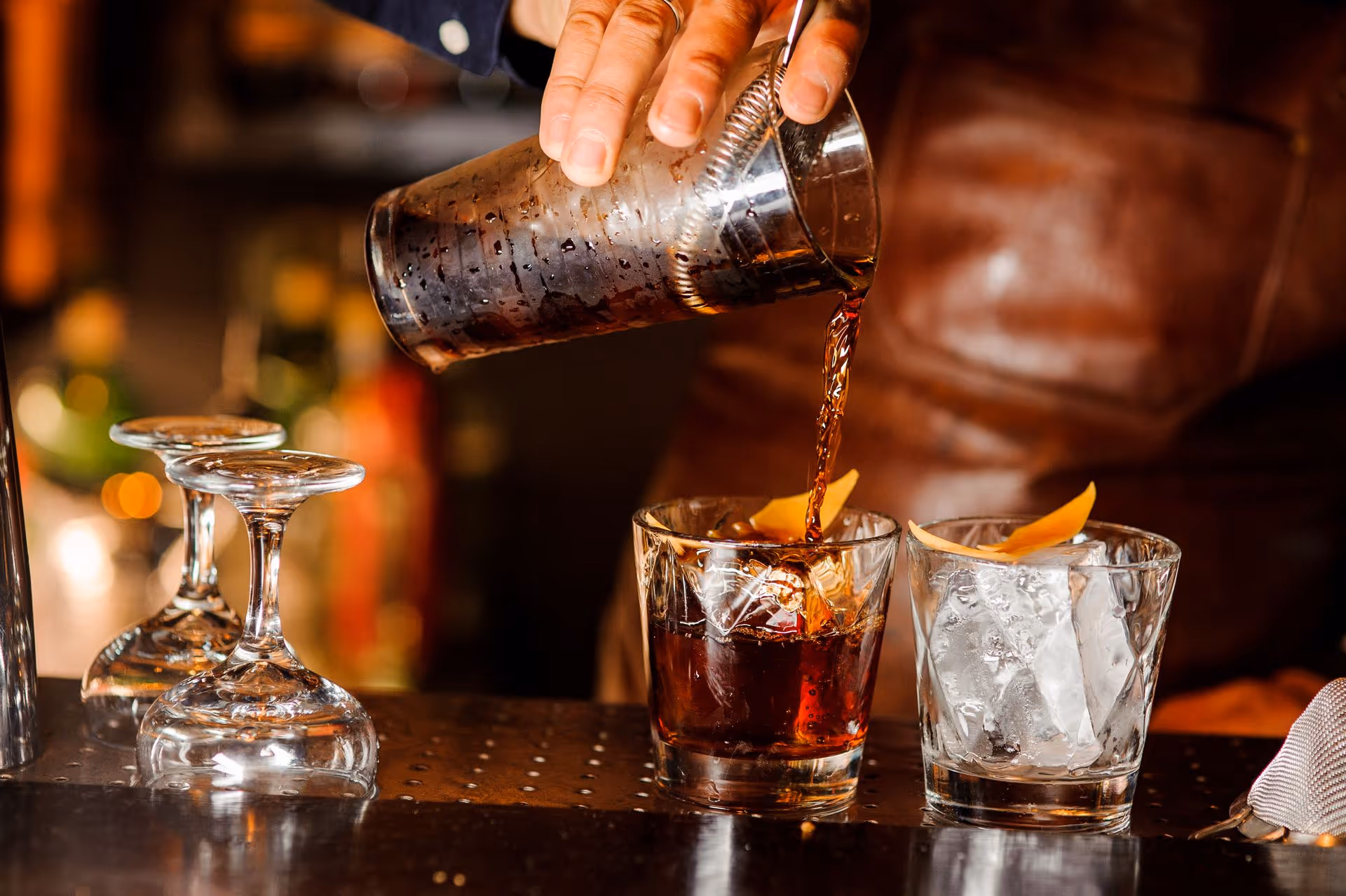 Bartender pouring dark cocktail from a shaker into a glass with ice and citrus peel, beside another glass on a bar counter. 