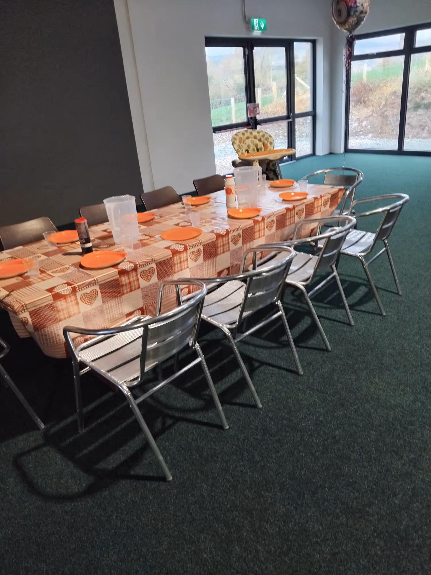 Dining table with orange plates and plastic cups, surrounded by chairs and a high chair.