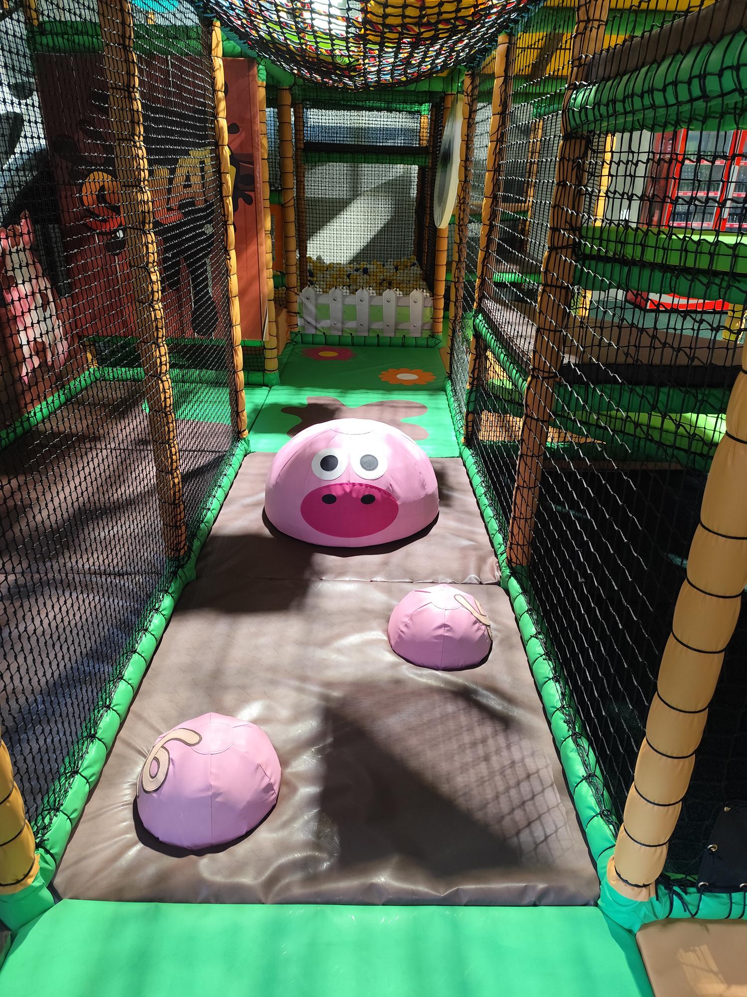 Indoor children's play area with a soft padded floor featuring three pink mound shapes, one with a smiling face resembling a pig.