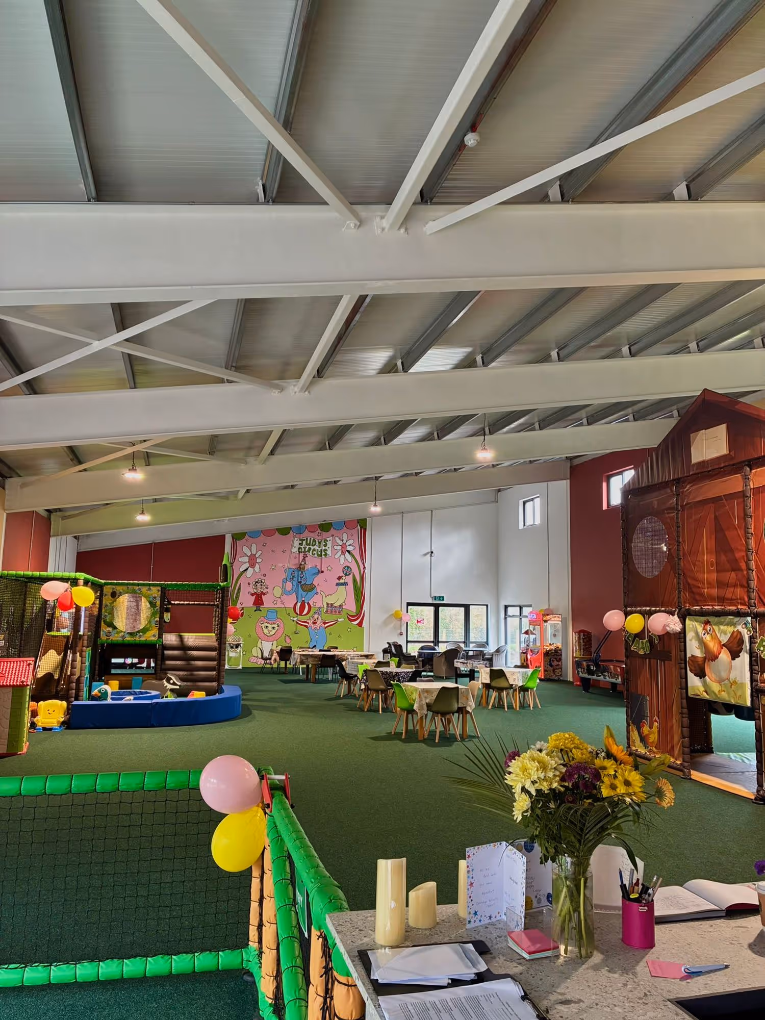 Indoor children's play area with green carpet, play structures, small tables and chairs, colorful balloons, and a floral bouquet on a counter.