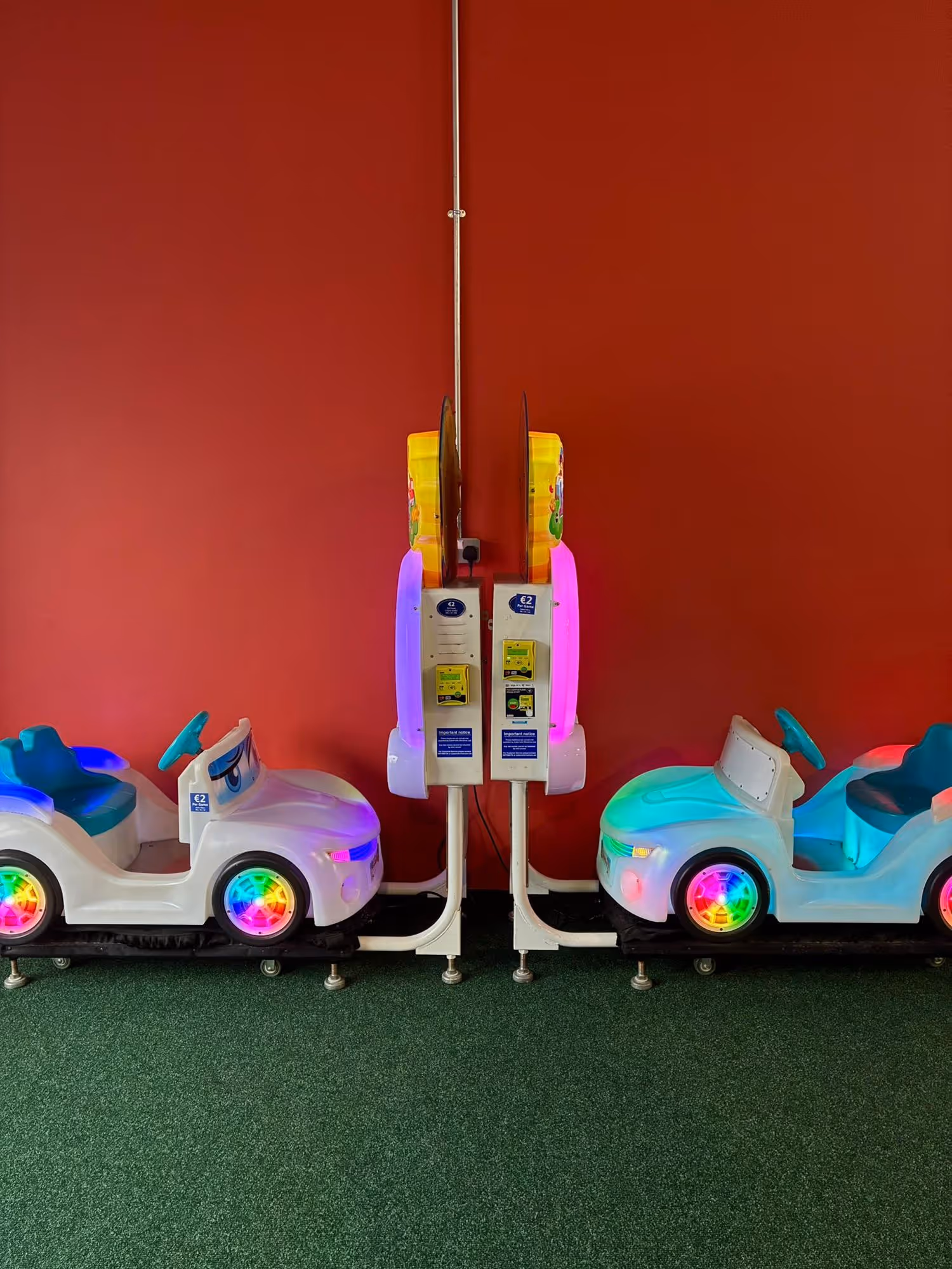 Two white children's ride-on cars with colourful illuminated wheels and hooked up to an arcade screen, both facing each other.