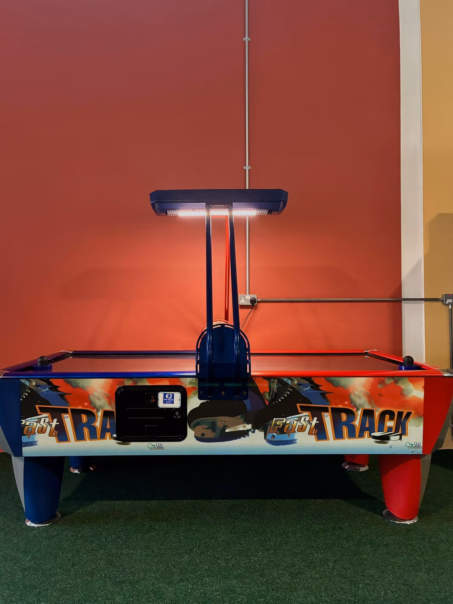 Air hockey table with blue and red sides under a hanging light against a red wall.