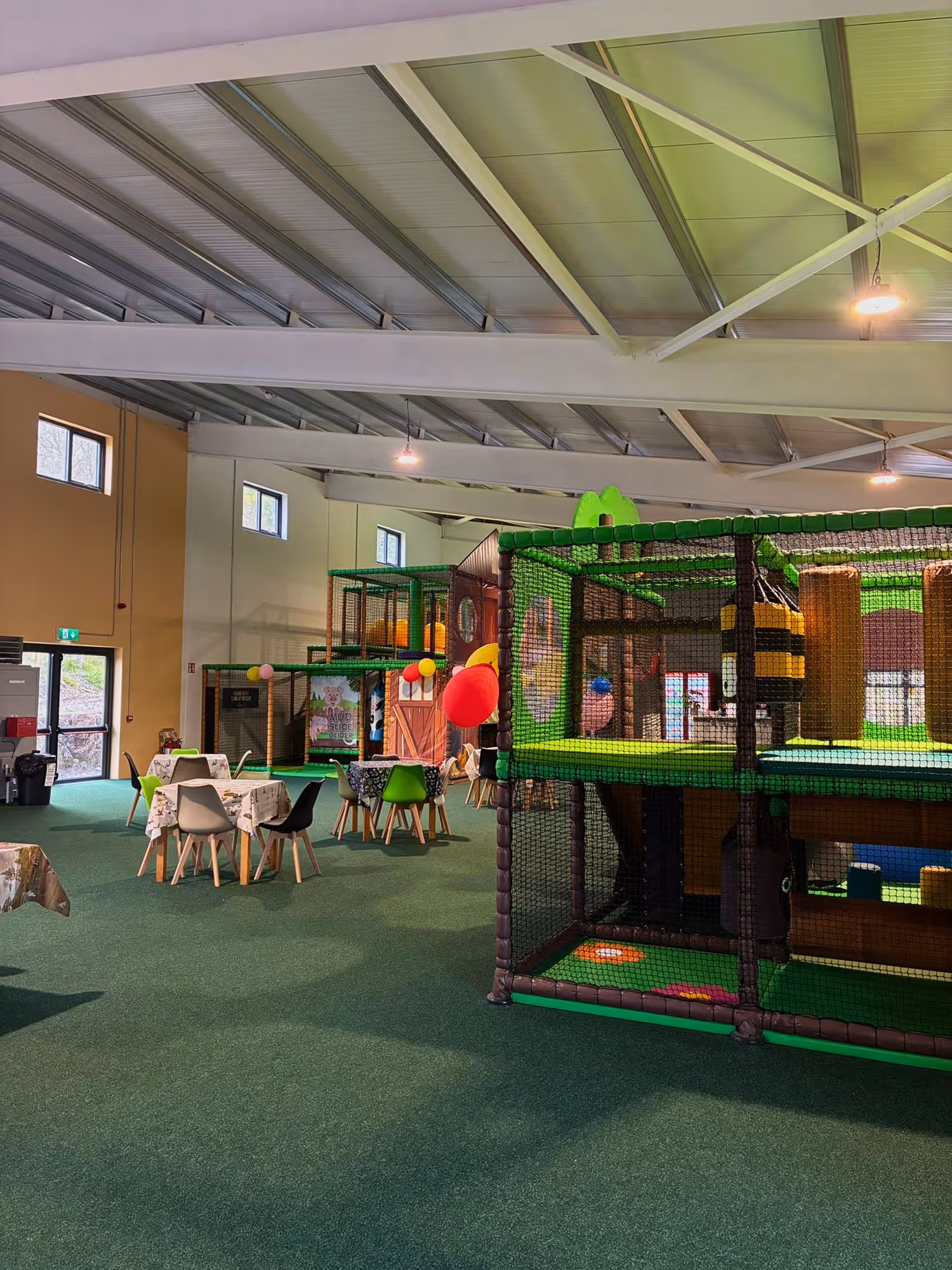 Indoor children's play area with soft play structures, tables with chairs, and green carpeted floor.