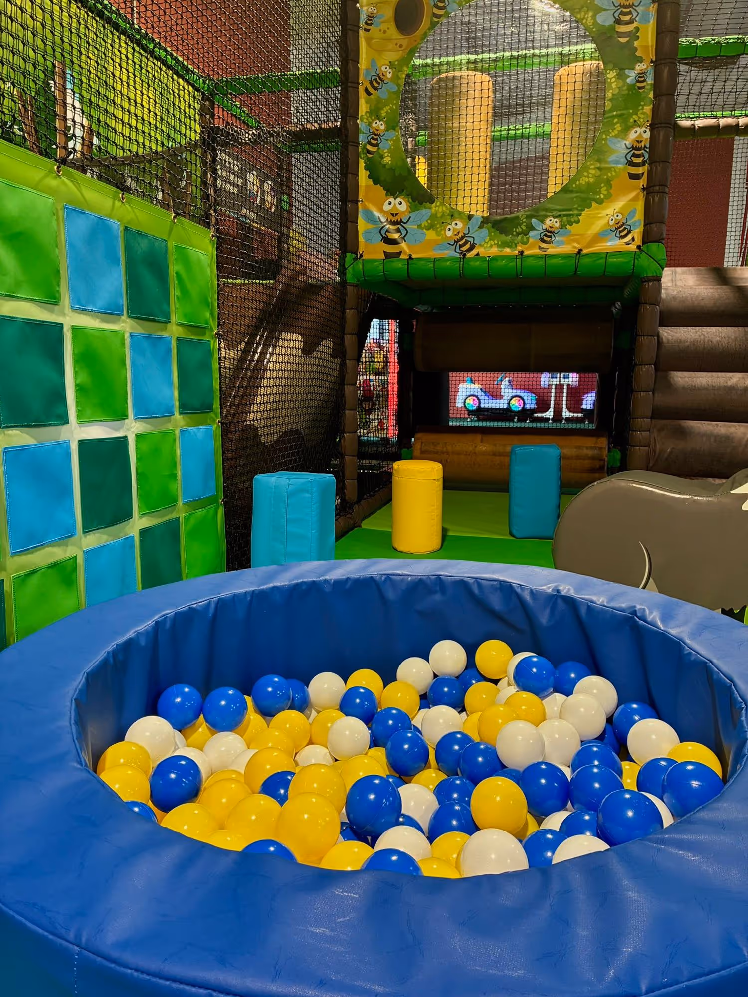 Indoor children's play area featuring a blue padded ball pit filled with yellow, blue, and white balls, netted climbing structures, and soft play obstacles.