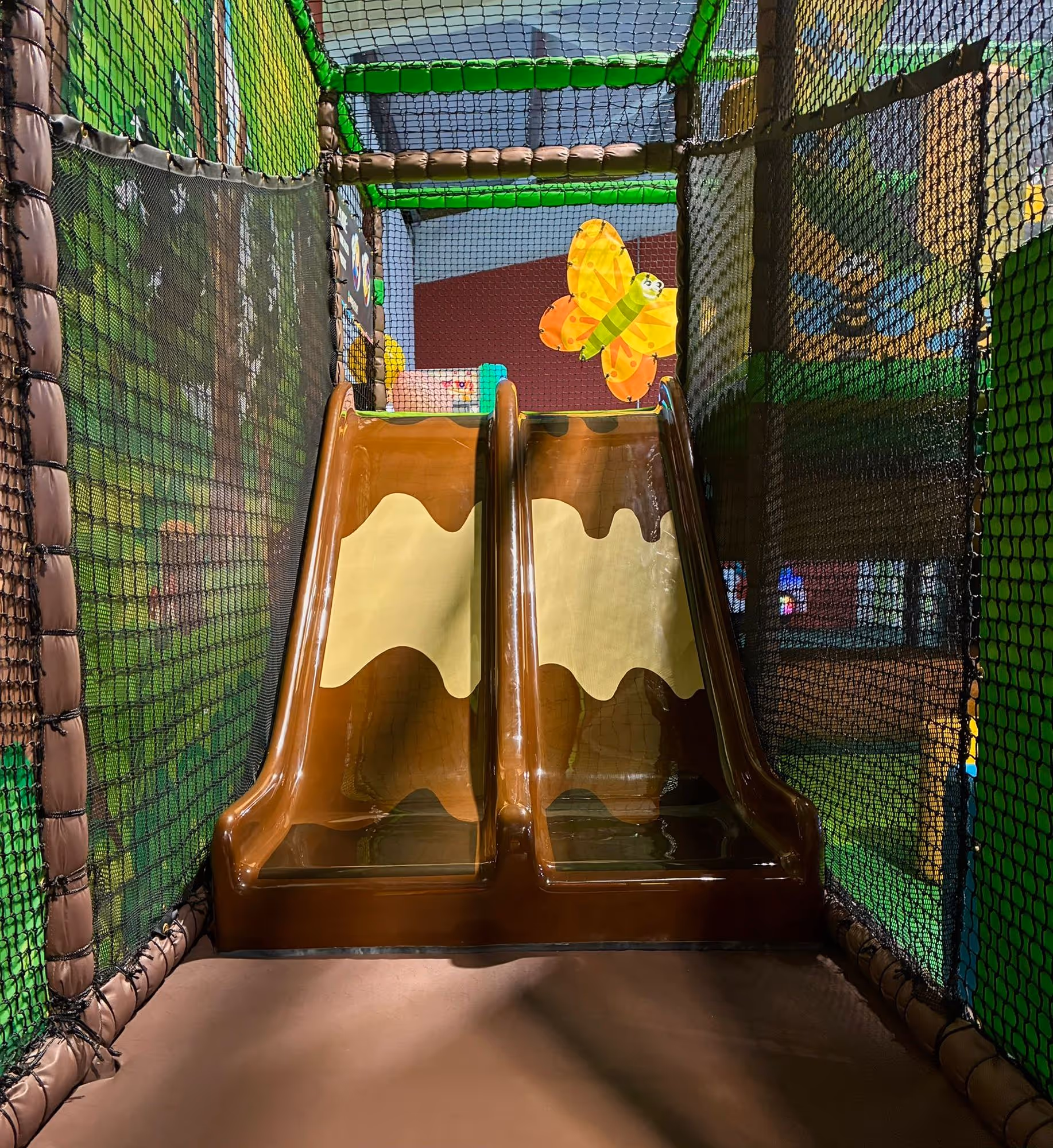 Indoor children's play area featuring brown double slides with a dripping pattern and safety netting on the sides.